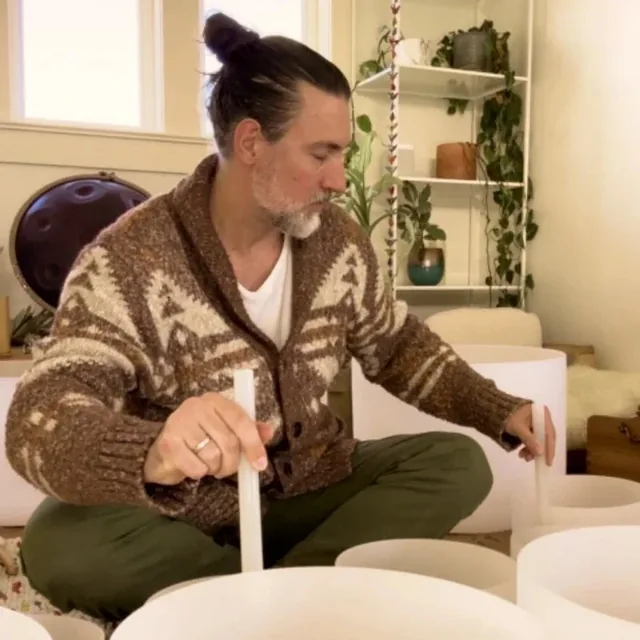 Graham sitting cross-legged among crystal singing bowls, holding a striker