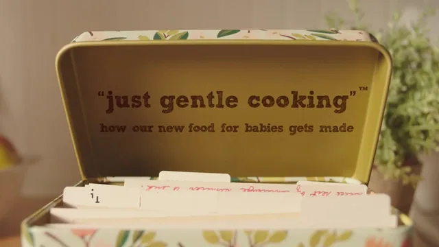 An open recipe tin on a kitchen counter, the inside of its lid reading “just gentle cooking,” how our new food for babies gets made, above a row of recipe cards