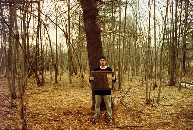 A man stands among bare trees holding an empty frame