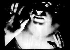 A face behind a raised hand on a television screen, in stark black and white