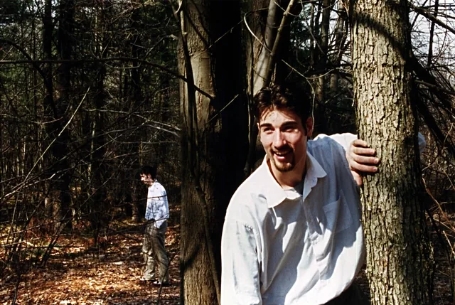 A man in a white shirt calls out beside a tree, another figure behind him