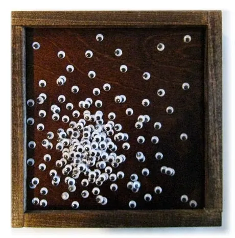 A spray of white googly eyes on dark wood, in a frame