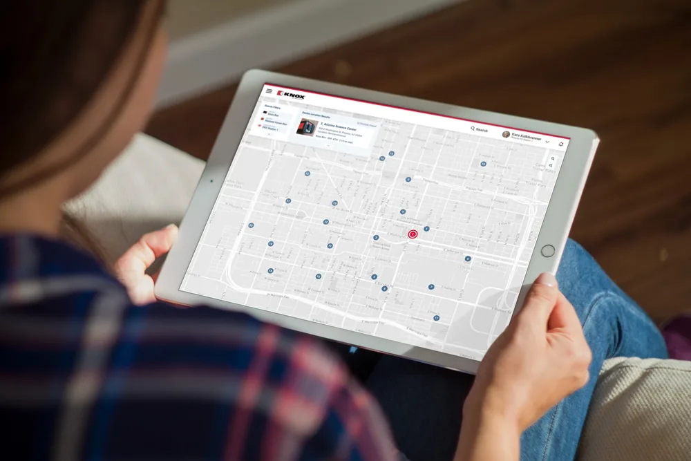 A person in a plaid shirt holds a tablet showing the Knox device map: numbered device pins across downtown Phoenix, search filters and a result card for a Knox Box at the Arizona Science Center