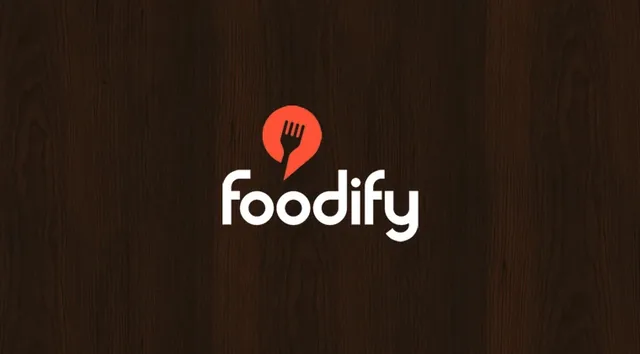 The Foodify logo in white with a red fork pin, on dark wood