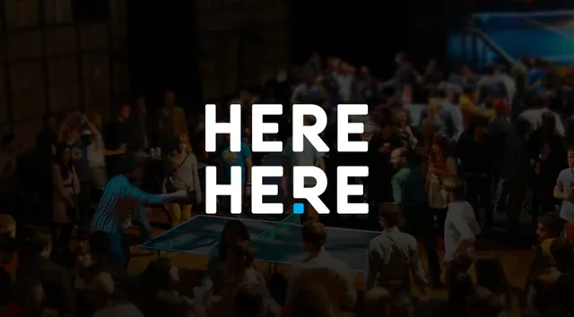 The Here Here logo: HERE HERE in white capitals over a dim crowd around a ping pong table, with a blue square before the last R